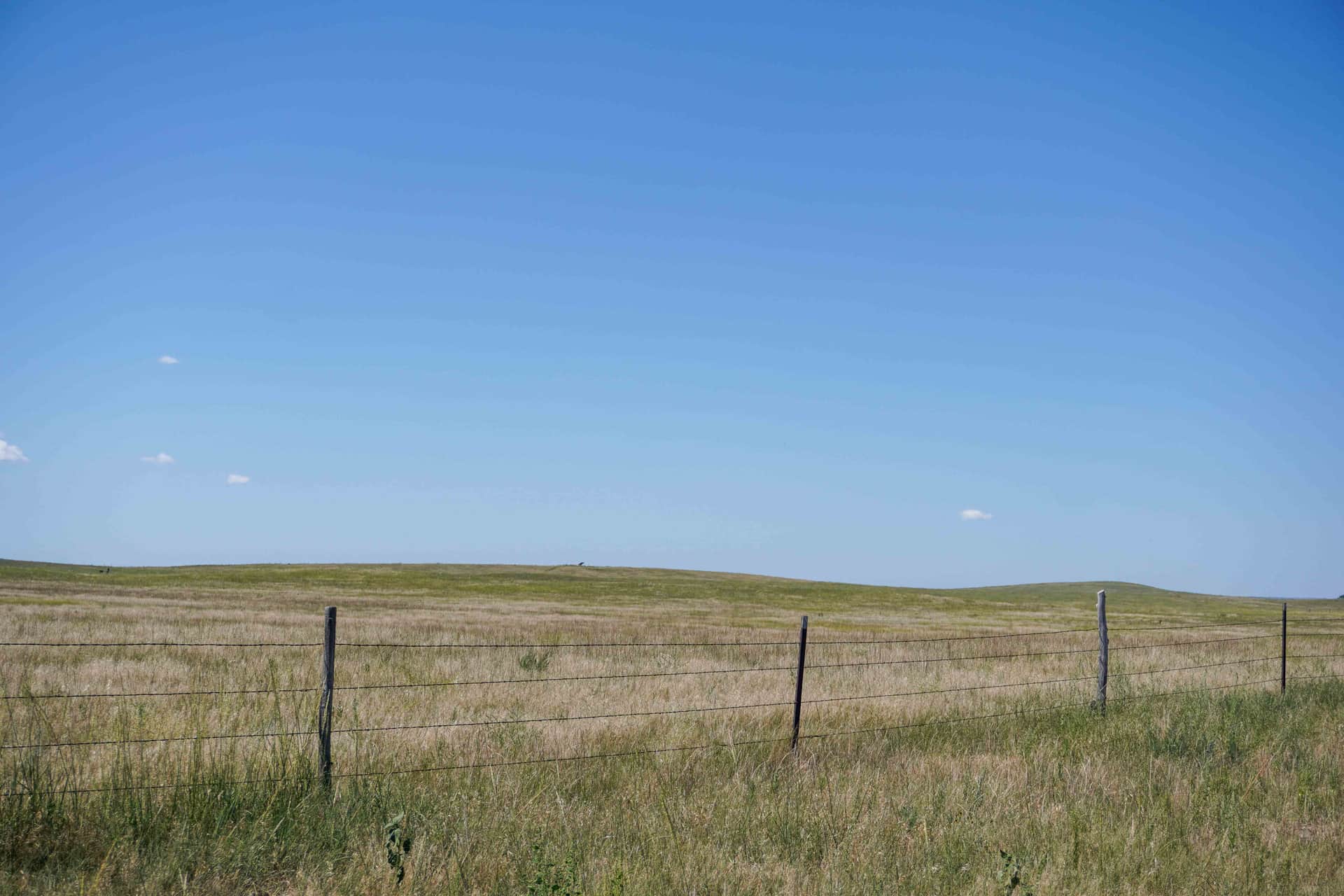 A barbed-wire fence line running across open Nebraska grassland prairie under a big blue sky, with low rolling hills on the horizon.