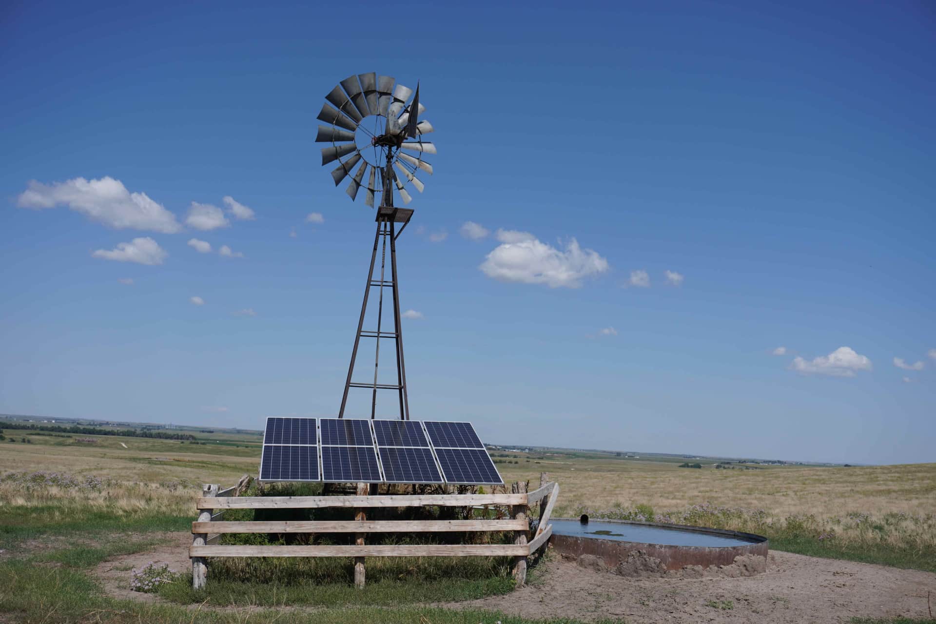 A classic multi-blade windmill on a lattice tower beside solar panels and a round steel stock tank on open Nebraska Sandhills range under a big blue sky.